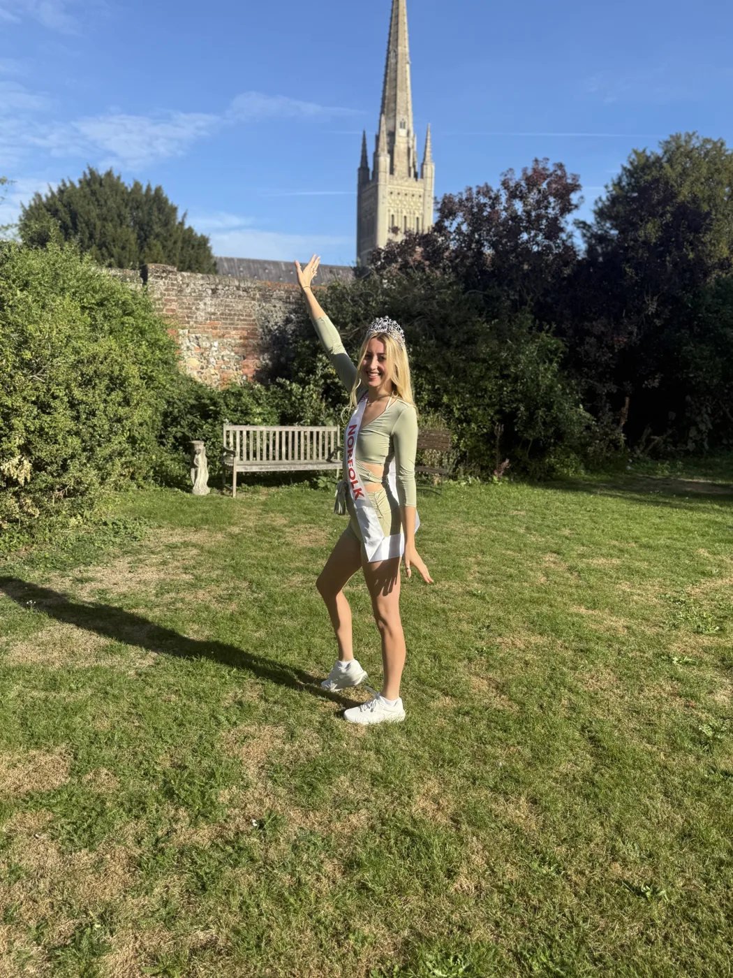 Amy Morris in a crown and Norfolk sash pointing up at a tall stone spire behind a garden wall